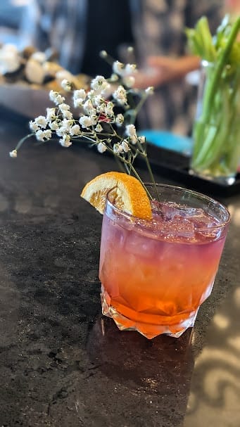 A close up of a coral-colored cocktail with an orange slice and baby's breath garnish on the Benson Theatre bar counter